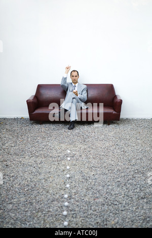 Businessman sitting on sofa dans le gravier, de lancer des balles de golf, pleine longueur Banque D'Images