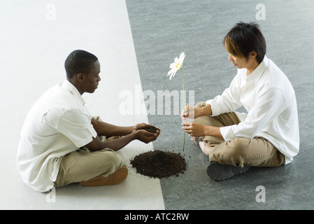 Deux hommes assis face à face sur le sol, un holding out poignée de terre, l'autre tenant une fleur Banque D'Images