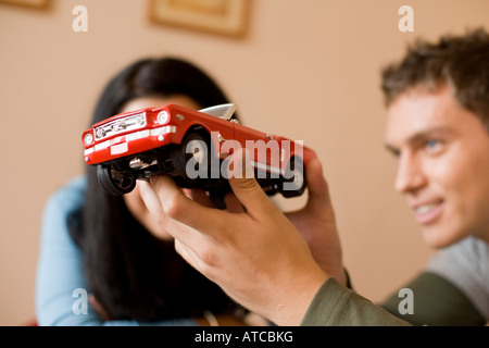 Jeune homme tenant près de son visage une petite voiture rouge de l'analyse et de jouer avec elle et une femme debout près de lui Banque D'Images
