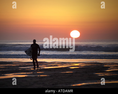 Silhouette d'un surfeur marche vers le haut de la plage au coucher du soleil. Widemouth Sands / Widemouth Bay, Bude, Cornwall, UK, British Isles Banque D'Images