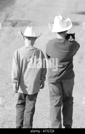 Deux garçons avec des chapeaux de cow-boy marchant sur un chemin vicinal avec chat sur l'épaule des garçons Banque D'Images