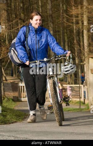 Jeune femme souriante poussant son vélo dans le chemin d'Nant yr Arian parc forestier de Galles Ceredigion après une balade dans les bois Banque D'Images