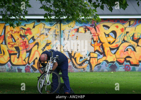 Une tête chauve homme habillé en bleu ajuste son vélo dans un parc de Londres devant un mur couvert de graffitis Banque D'Images