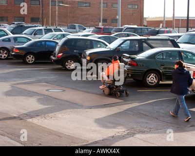 Un homme handicapé dans un fauteuil roulant motorisé transit par un parking de l'USA. Banque D'Images