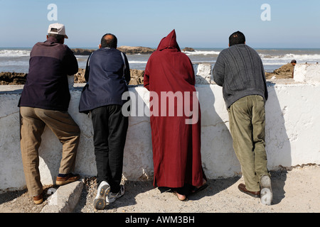 Quatre hommes se penchent contre un mur qui donne sur la mer, trois d'entre eux portant des vêtements modernes et un portant des vêtements traditionnels, es Banque D'Images