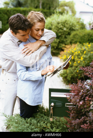 Woman holding mail, l'homme l'étreindre par derrière, à côté de la boîte de courrier électronique, des arbustes en arrière-plan Banque D'Images