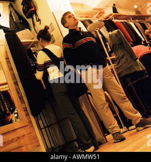 Jeune femme à la recherche de vêtements dans une boutique de vêtements, et young man standing with hands in pockets Banque D'Images