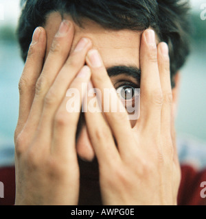 Jeune homme avec les mains sur le visage, regardant à travers les doigts avec un oeil, close-up Banque D'Images