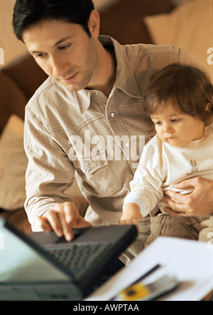 Le père et le bébé, le père à l'aide d'ordinateur portable, bébé assis sur les genoux du père. Banque D'Images