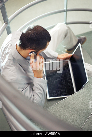 Homme assis dans les escaliers avec un téléphone mobile et un ordinateur portable, high angle view Banque D'Images