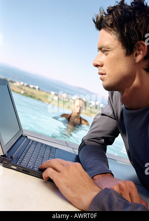 Man using laptop computer, woman in swimming pool in background Banque D'Images