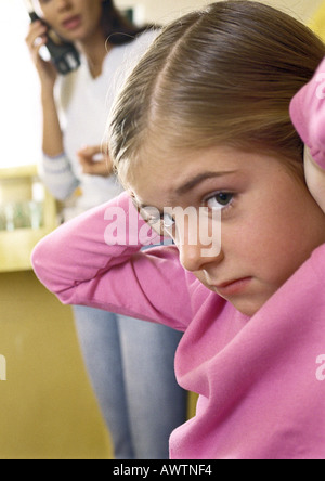 Petite fille avec les mains sur les oreilles, looking at camera, close-up, Woman talking on phone in background Banque D'Images