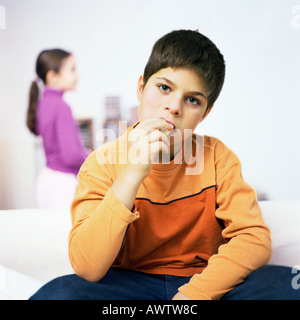Garçon assis sur le canapé, manger, Girl standing in background Banque D'Images