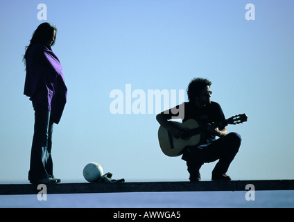 Femme debout, homme accroupi, jouer de la guitare sur un banc Banque D'Images