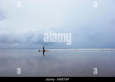 Lone surfer assis sur une planche de surf dans une eau peu profonde, side view Banque D'Images