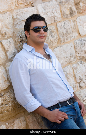 Jeune homme portant des lunettes de soleil à côté d'un mur, les mains sur son pantalon Banque D'Images