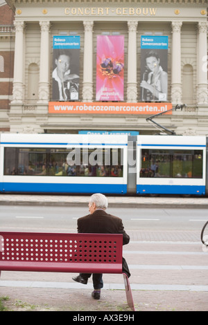 Vieil homme assis sur un banc en face d'une salle de concert de l'autre côté de la rue, tandis que le tramway passe par Banque D'Images