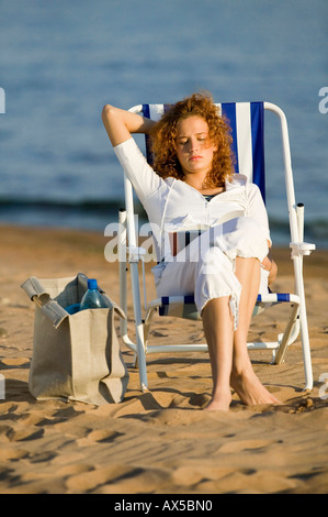 Jeune femme assise dans une chaise longue sur la plage, reading book Banque D'Images