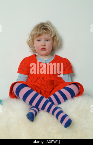 Petite fille blonde assis les jambes croisées sur un tapis de fourrure, en robe rouge et de collants à rayures. Banque D'Images