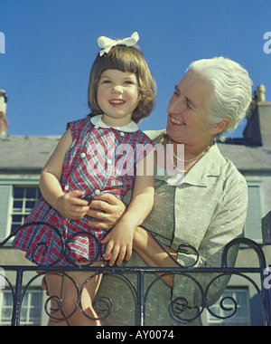 Sourire fier grand-mère aux cheveux gris debout et tenant sa petite-fille, près du haut de la porte de jardin en fer forgé l'Angleterre Banque D'Images