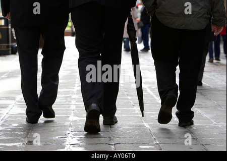 Trois hommes d'affaires en costumes sombres avec un parapluie marche sur chaussée mouillée dans la zone piétonne du centre-ville de Belfast Banque D'Images