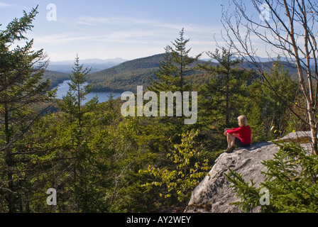 Une femme dans un tee shirt rouge assis sur une saillie rocheuse au sommet le Mont Owl's Head en regardant le lac et les montagnes au loin. Banque D'Images