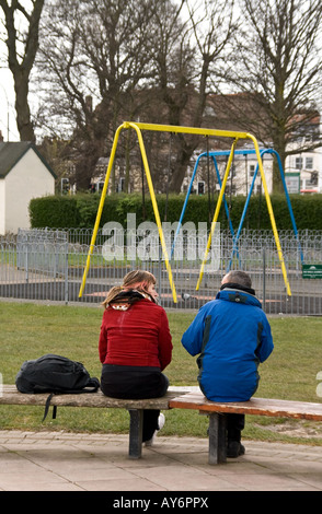 L'homme et la femme assis sur un banc de parc avec dos à nous, de parler, avec des balançoires en face d'eux Banque D'Images