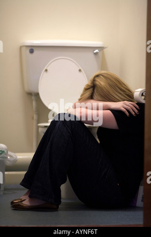 Close up d'armes et les épaules de l'adolescent aux cheveux blonds femme assise en position fœtale sur le sol de toilettes au Royaume-Uni Banque D'Images