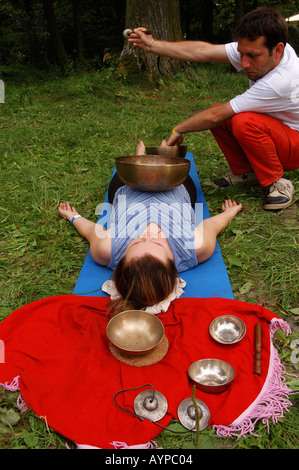 Un homme effectuant la guérison de massage sonore tibétain sur une femme allongée sur un tapis à l'extérieur, en utilisant des bols chantants pour la thérapie dans un cadre de parc naturel. Banque D'Images