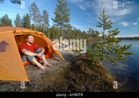 Homme assis en face d'une tente à un lac, à distance, dans le Parc National, Femundsmark Femundsmarka, Norvège Banque D'Images