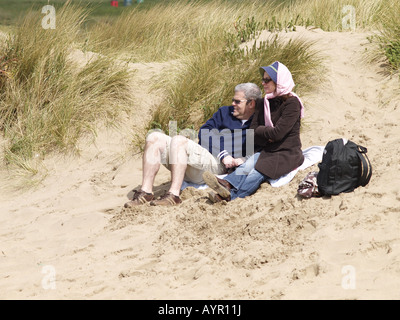 Vieux couple sitting on sand dunes à Summerleaze Beach, Cornwall, sur une journée de printemps venteux. Banque D'Images