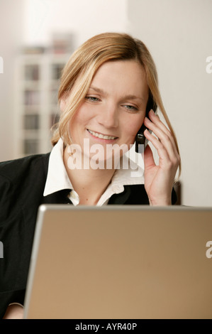 Young blonde woman in front of laptop, talking on phone, smiling Banque D'Images