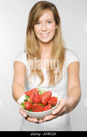 Jeune femme avec de longs cheveux blond foncé offrant un bol de fraises fraîches Banque D'Images