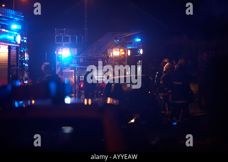 Les pompiers en attente derrière un camion d'incendie moteur de pompage par des voitures en stationnement sur les lieux de l'incendie d'une maison au Royaume-Uni Banque D'Images