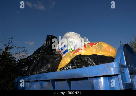 Commercial bleu wheely bin débordant de sacs en plastique d'ordures assis dans une aire rurale avec un ciel bleu clair 2/3 Banque D'Images