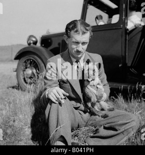 OLD VINTAGE SNAPSHOT DE L'HOMME ASSIS DANS LA ZONE HOLDING PETIT CHIEN AVEC VINTAGE CAR EN ARRIÈRE-PLAN Banque D'Images