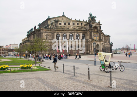 Opéra Semper à Dresde, en Allemagne, avec un jardin fleuri et un pavé au premier plan, un pousse-pousse dans la rue et des gens marchant Banque D'Images