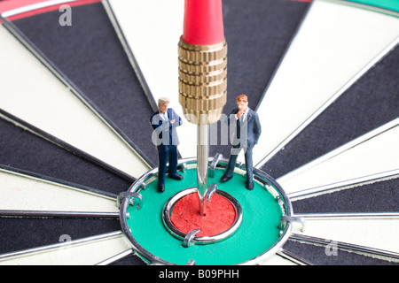 Businessman figurines debout sur un jeu de fléchettes avec une fléchette coincé dans l'oeil de taureaux Banque D'Images
