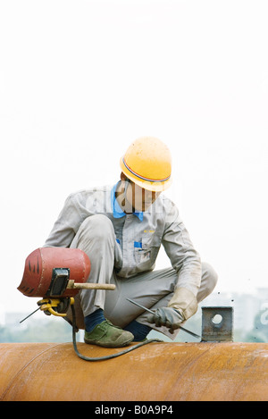 Worker crouching sur tuyau métallique, holding masque de soudage, regardant vers le bas Banque D'Images