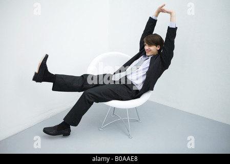 Young man sitting in chair, les jambes tendus, bras levés, pleine longueur Banque D'Images