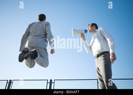 Businessman debout à côté de collègue, criant dans un mégaphone, collègue de sauter dans l'air, low angle view Banque D'Images