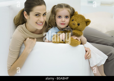 Jeune mère assise avec sa fille sur le canapé, little girl holding teddy bear, smiling at camera Banque D'Images