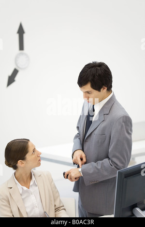 Surveillant masculin debout à côté d'employée, pointant montre-bracelet, aiguilles d'horloge sur le mur en arrière-plan Banque D'Images