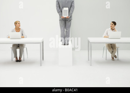 Deux femmes assis à un bureau avec les ordinateurs portables, les deux regardant man holding document behind back Banque D'Images