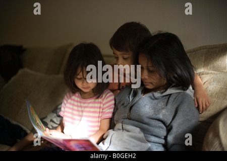 Mixed Race family mère et deux enfants lire un magazine pour enfants, assis sur un canapé dans un rayon de soleil Banque D'Images
