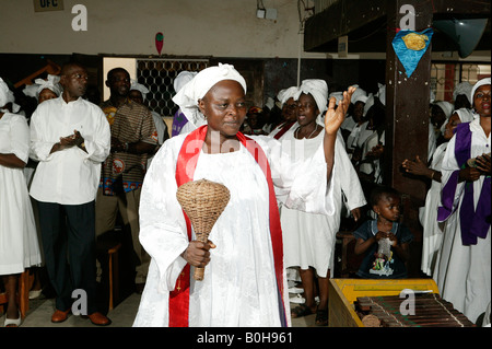 Femme vêtue de robes blanches et red sash de faire de la musique avec un hochet en osier pendant un service religieux à Douala, Cameroun, Afrique Banque D'Images