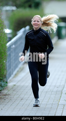 Les jeunes, smiling blonde woman jogging survêtement noir on sidewalk Banque D'Images