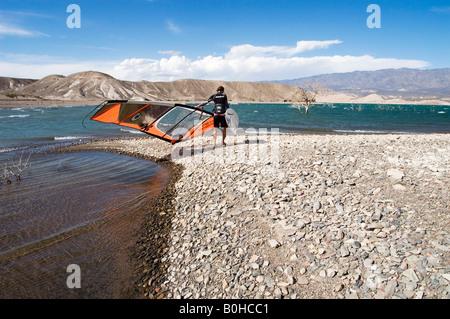 Windsurfer en combinaison avec son conseil et naviguer à travers le gravier arides balayées par le bord du lac entouré de montagnes, Cuesta d Banque D'Images