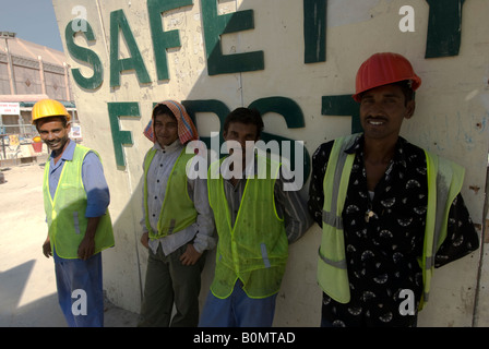 Quatre travailleurs migrants prendre une pause à l'ombre contre un mur où est écrit : La sécurité d'abord, Dubaï, Emirats Arabes Unis. Banque D'Images