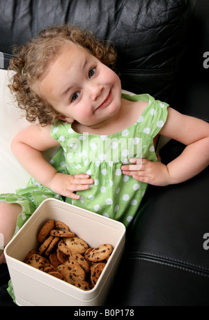 Une fillette de 3 ans assis sur le canapé à la maison avec une boîte de cookies sur ses genoux en mettant ses mains sur ses hanches Banque D'Images